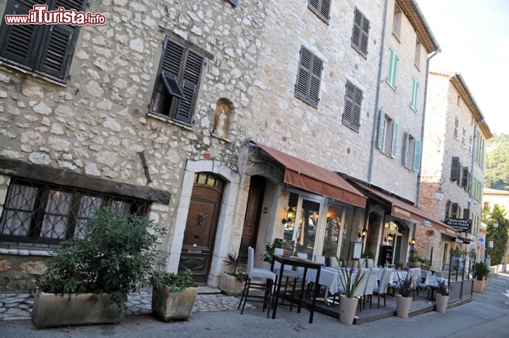 Immagine Ristorante a Tourrettes sur Loup, Francia - Sulla piazza principale del paese, Place de la Libération, si affacciano bar e ristoranti che durante quasi tutto l'anno, complici anche le temperture miti, permettono alla clientela di sedersi comodamente nei dehors esterni per assaporare i piatti della tradizionale cucina francese. In quest'immagine, un grazioso ristorante con i suoi tavoli sul lato che costeggia l'ingresso al parcheggio di Tourrettes sur Loup  © Sonja Vietto Ramus