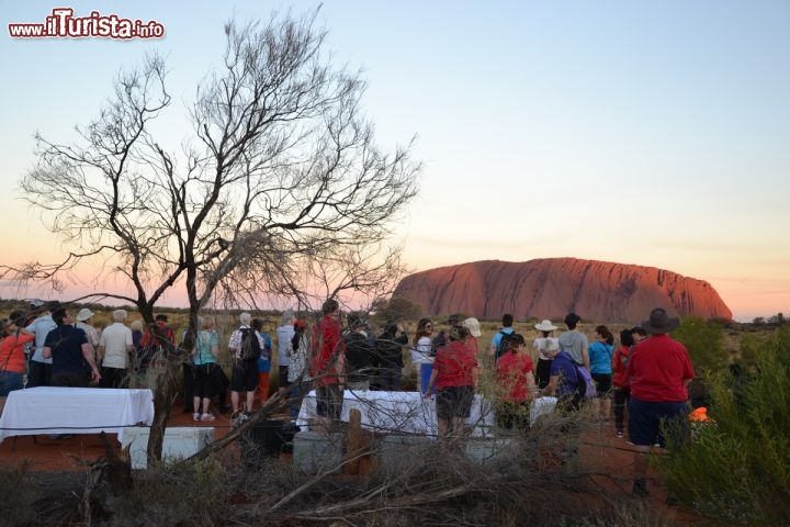 Immagine Turisti contemplano al tramonto Ayers Rock in Australia - Oltre al tradizionale aperitivo al tramonto, una delle esperienze più belle che si possa compiere ad Uluru è la cena denominata come "Sound of Silence experience" cioè l'esperienza del suono del silenzio. Il silenzio è quello dell'outback australiano, che qui consente di vivere la magia ancestrale della notte, tra stelle e via lattea come mai le avete vissute prima, e la sagoma della montagna sacra che renderà indimenticabile la vostra esperienza, lontani dai rumori e le luci della "civiltà".