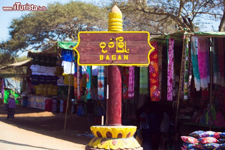 Immagine Cartello stradale per Bagan di fronte a un mercato, Myanmar. Stoffe colorate in una bancarella al mercato fanno da sfondo a questo caratteristico cartello di segnalazione stradale che indica la città di Bagan - © dmitry_islentev / Shutterstock.com