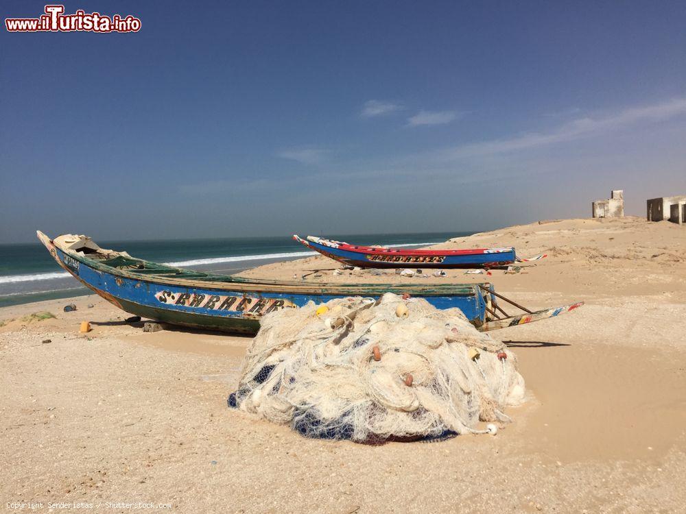 Immagine Due tradizionali barche in legno sul litorale nei pressi di Nouakchott, Mauritania. In primo piano, reti da pesca e sullo sfondo alcune abitazioni - © Senderistas / Shutterstock.com