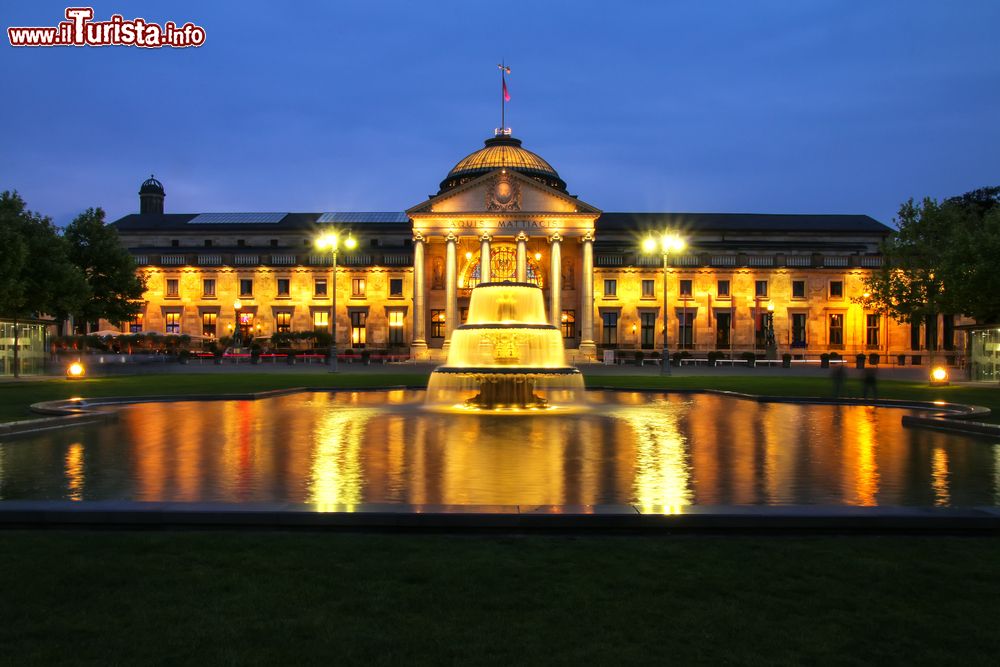 Immagine Il Kurhaus e il Bowling Green illuminati di sera, Wiesbaden, Germania. Il Bowling Green è un rettangolo allungato con prati e due piscine: il termine Bowling Green riporta al gioco con la palla, in inglese "bowls". All'inizio del XX secolo, l'area verde era chiamata Kursaalplatz.