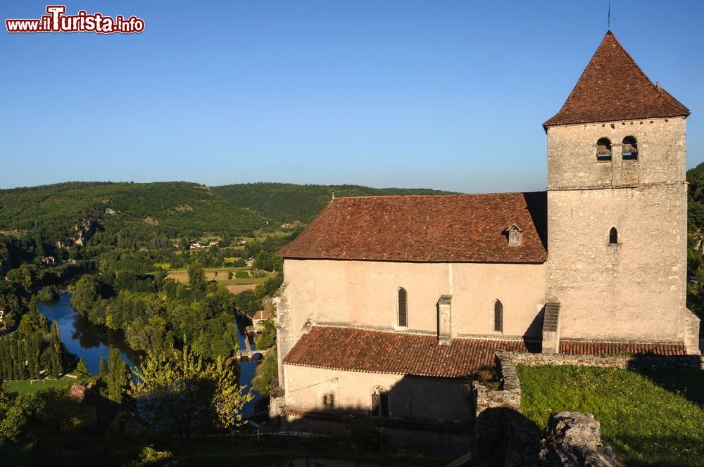 Immagine La chiesa di Saint-Cyr e Sainte-Julitte a Saint-Cirq-Lapopie, Occitania (Francia). Su un lato, il fiume Lot circondato da una vegetazione lussureggiante.