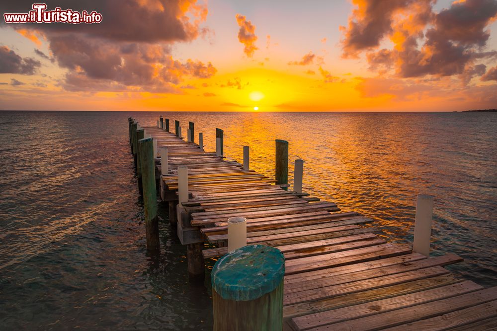 Immagine Un molo in legno fotografato durante il tramonto sull'isola di Eleuthera, Bahamas. I riflessi dorati del cielo si rispecchiano sull'acqua del mare e sulla passerella dove attraccano le barche dei pescatori.