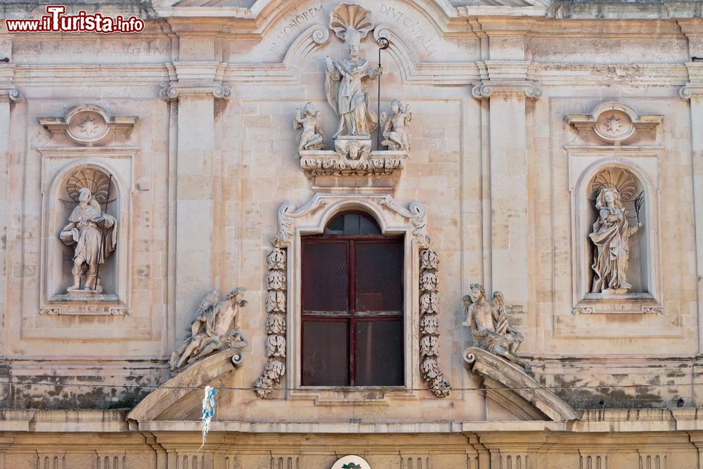 Immagine Parte alta della facciata del duomo di San Cataldo, Taranto, Puglia. Tagliata orizzontalmente da un architrave spezzato in stile barocco, la facciata si presenta con due nicchie con angeli che guardano verso il rettangolo del finestrone centrale in cui si trova adagiata la statua di San Cataldo.