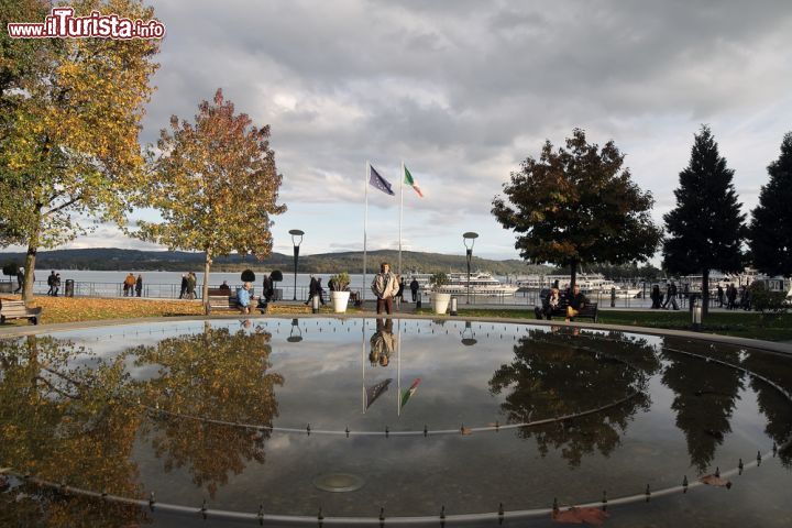 Immagine Passeggiata lungo il lago Maggiore a Arona, Piemonte - Considerato il più esteso dei laghi prealpini, tanto da meritarsi il nome di "Maggiore", questo bel lago ha incantato grandi artisti italiani e stranieri che hanno soggiornato proprio sulle sue sponde. Per scoprirne gli scorci paesaggistici più suggestivi si può passeggiare lungo le sue rive ammirando castelli e ville, giardini botanici e riserve naturali. In questa immagine una bella fontana a forma circolare in cui svettano le bandiere italiana e dell'Unione Europea © marcovarro / Shutterstock.com