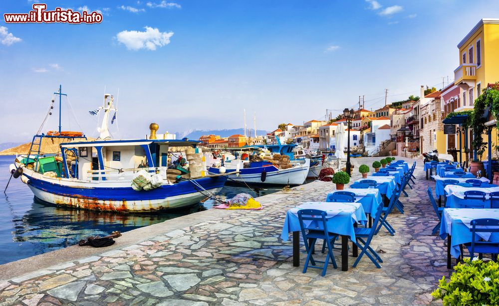 Immagine Uno scorcio del porto sull'isola di Chalki, Grecia, con barche ormeggiate e taverne con tavoli e sedie.