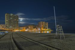 La spiaggia di Oropesa del Mar fotografata di notte, Spagna. Le luci dei palazzi che si affacciano sul lungomare si riflettono nelle acque della baia e rischiarano la fine sabbia dorata delle ...